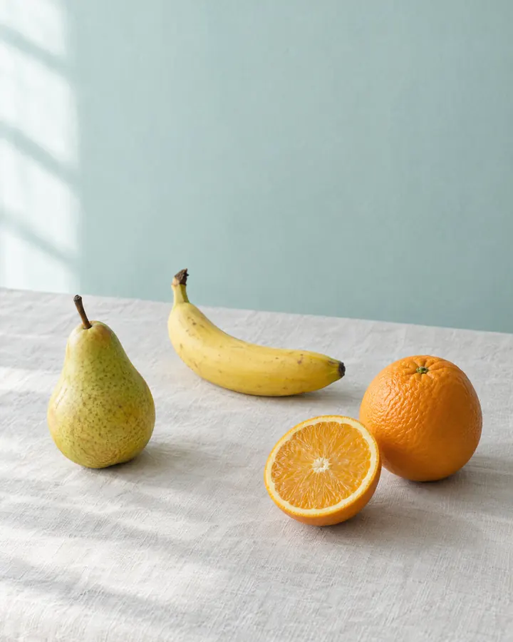 Pera, banana e laranja sobre uma mesa clara.